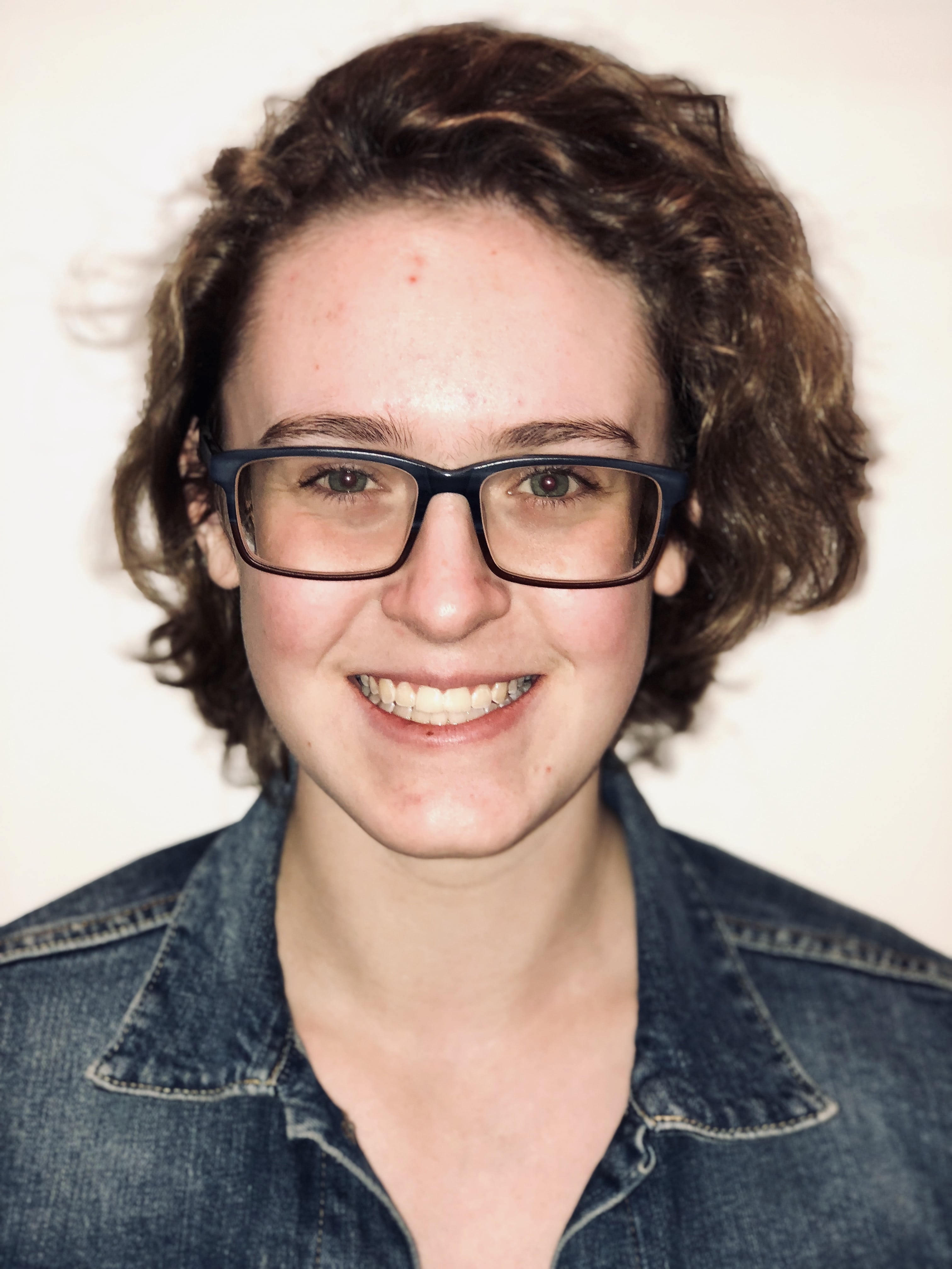 A head and shoulders picture of a smiling young woman in a jean jacket with short, curly brown hair, green eyes and glasses.